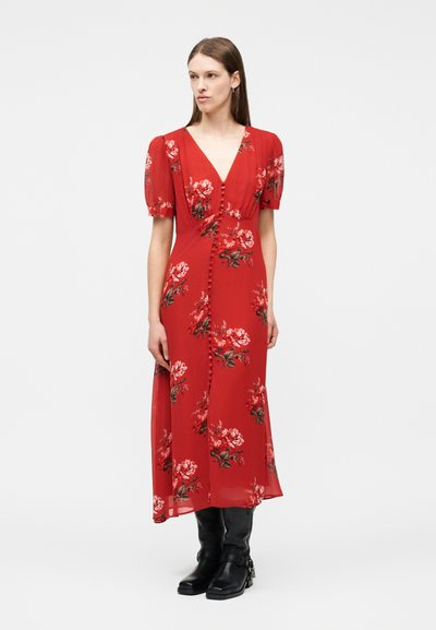 Naine seisab ja kannab punast lillelist midi-kleidiga koos puff varrukatega ja mustade pahkluu saapadega, taustal on hele ühtlane taust.