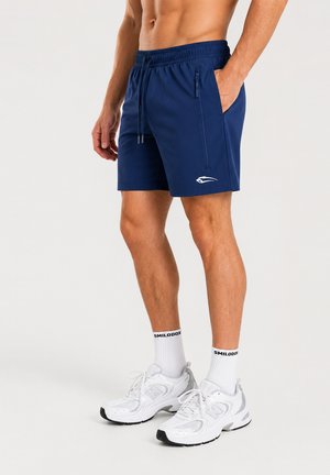 Homme portant un short de sport bleu marine avec des poches zippées, des chaussettes blanches montantes avec texte noir et des chaussures de course blanches sur fond uni.