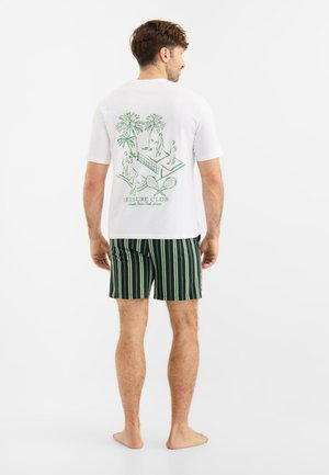 Mann steht barfuß, trägt ein weißes T-Shirt mit grünem Tennis-Motiv und schwarze Shorts mit grünen Streifen, ist von hinten zu sehen.