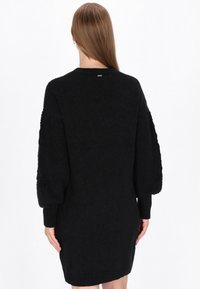 Sort strikket kjole med en tekstureret liv og voluminøse ærmer, afsluttet med en ribbet bund og diskret branding på ryggen.