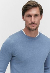 Ljusblå stickad tröja med rund halsringning, långa ärmar och slät textur. Bärs över en vit skjorta. Minimalistisk design, inga synliga mönster.