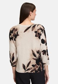 Femme portant un pull beige avec des motifs floraux bruns et noirs, à manches trois quarts, et un pantalon noir, de dos contre un fond uni.