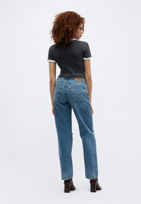 Γυναίκα με σγουρά μαλλιά, φοράει μαύρο στενό t-shirt, ψηλόμεσο μπλε τζιν και σκούρα μποτάκια με τακούνι, γυρισμένη προς τα πίσω.