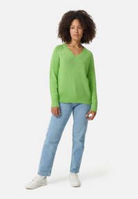 Groene gebreide v-hals trui met lange mouwen, gecombineerd met lichtblauwe jeans en witte sneakers. De trui heeft een geribbelde zoom en manchetten.