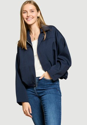 Jeune femme aux longs cheveux blonds sourit, portant une veste bleu marine par-dessus un haut blanc et un jean bleu, sur un fond clair uni.