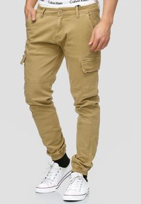 Beige cargo pants with elastic cuffs, multiple pockets, and a relaxed fit. Worn with white sneakers and a visible logo waistband.