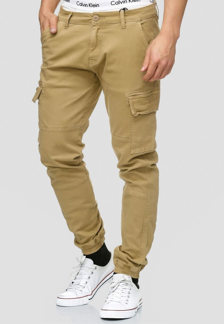Beige cargo pants with elastic cuffs, multiple pockets, and a relaxed fit. Worn with white sneakers and a visible logo waistband.