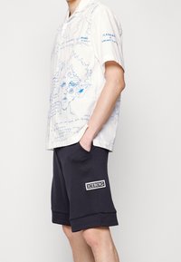 Biała koszula z krótkim rękawem z niebieskimi wzorami; zestawiona z granatowymi szortami z naszywką logo Iceberg po lewej stronie. Casualowy, luźny krój.