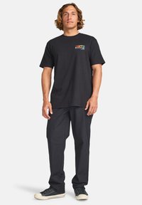 Black cotton t-shirt featuring a colorful graphic on the left chest, paired with loose-fitting black trousers and dark sneakers.