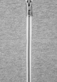 Felpa grigia con cerniera argentata, tessuto strutturato, cucitura centrale e cuciture piatte lungo i bordi.