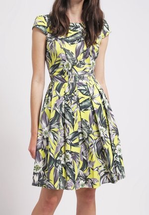 Femme portant une robe jaune à manches courtes avec un grand motif floral de feuilles vertes et blanches et une jupe plissée.