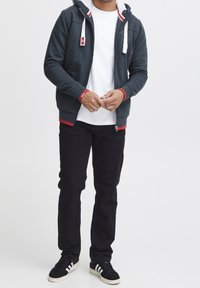 Sweat à capuche zippé gris foncé avec doublure en fourrure blanche et accents rouges, porté sur un t-shirt blanc, assorti à un jean noir et des baskets noires.