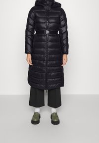 Černý péřový kabát s kapucí, s páskem v pase, horizontálně prošívaný design a plný zip. Nosič má na sobě široké černé kalhoty a zelené boty.