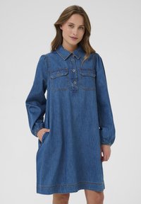 Džinsu kleita ar apkakli, garām pūkainām piedurknēm, divām krūšu kabatām un sānu kabatām; ar brīvu piegriezumu un redzamām stīgām.