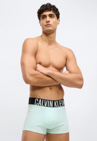 Kailis vīrietis ar tumšiem, čokurotiem matiem stāv ar rokām saslēgtām, uzvilcis gaiši zilos Calvin Klein bokseršortus pret vienkāršu baltu fonu.
