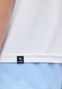 Τμήμα ενός λευκού πουκάμισου με μαύρη ετικέτα UPF50 πάνω από ανοιχτό μπλε παντελόνια, που δείχνει μέρος του χεριού ενός ατόμου.