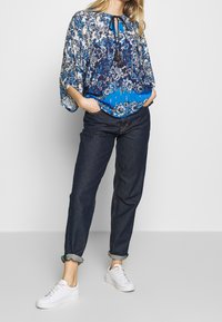 Kvinna iklädd blommönstrad blus i blått med tofsar, mörka uppvikta jeans och vita sneakers som står mot en ljusgrå bakgrund.