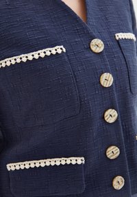 Veste texturée bleu marine avec quatre gros boutons, deux poches bordées de crème et un col en V. Tissu doux avec un motif subtil.
