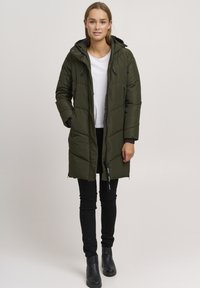 Olivengrønn puffjakke med hette, quiltet design, sidelommer og svarte detaljer. Brukt med en hvit skjorte og svarte bukser.