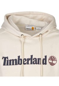 Haut à capuche couleur crème avec des cordons de serrage et une poche kangourou. Présente le logo "Timberland" en brun foncé et une icône d'arbre. Texture douce.