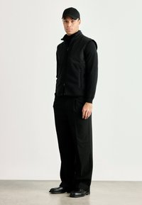 Colete preto sem mangas sobre uma blusa preta de manga longa, combinado com calças pretas. O conjunto apresenta um design minimalista e uma textura suave.