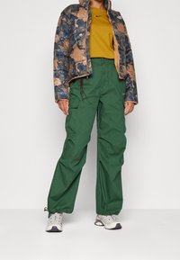 Puffjakke med blå og brun camouflage mønster, grønne cargo bukser og grå sneakers med lilla accenter.