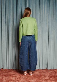 Mulher com cabelo comprido, vestindo uma jaqueta verde curta, calças azuis largas com pontos brancos e saltos brancos, em pé em frente a cortinas azuis.