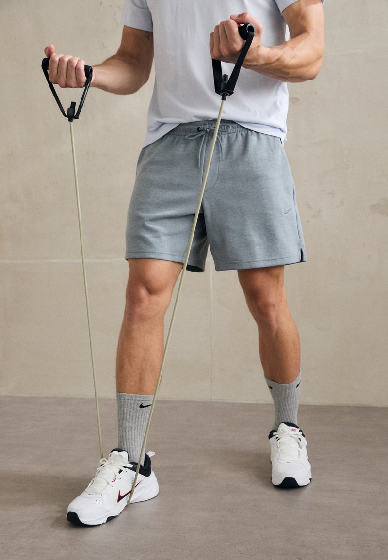 Benzile de rezistență sunt ținute în mâinile unei persoane care poartă un tricou gri deschis, pantaloni scurți gri și adidași albi cu accente roșii. Șosete gri sunt vizibile.
