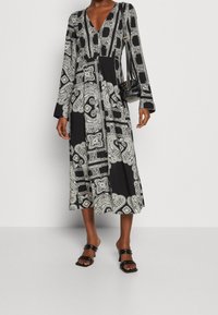 Robe noire à manches longues avec des motifs paisley et géométriques blancs, décolleté en V, silhouette fluide, assortie à un sac à main noir et des sandales.