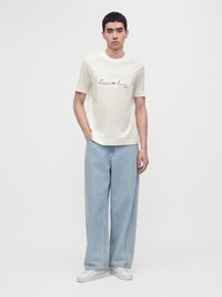 Νέος άνδρας φορά ένα λευκό t-shirt Emporio Armani, ανοιχτό μπλε τζιν παντελόνια με φαρδιά μπατζάκια και λευκά αθλητικά παπούτσια, στέκεται μπροστά σε λευκό φόντο.