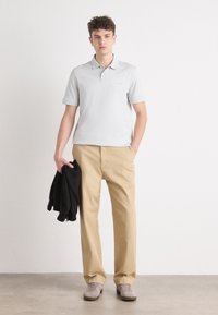 Polo cinza claro, calças bege, segurando uma jaqueta preta, com sapatilhas cinzas. Design simples, mangas curtas e corte descontraído.