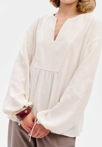 Langärmlige, cremefarbene Bluse mit V-Ausschnitt, geraffter Taille und bestickten Mustern an den Ärmeln, locker sitzend und mit weicher Textur.