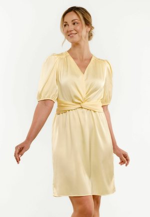Smiling woman in a pale yellow satin dress with puffed short sleeves and a twisted knot waist detail, posing against a white background.