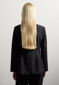 Persona con pelo rubio largo y lacio, vistiendo una chaqueta negra y pantalones negros, de pie frente a una pared gris claro y lisa, mirando hacia el otro lado.