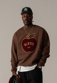 Hnedé mikiny s textúrovaným motívom jablka so žltou a červenou výšivkou, v kombinácii s bielou košeľou a tmavými rifľami.