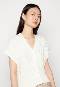 Weißer kurzer Cardigan mit einem Kabelstrickdesign, kurzen Ärmeln und einem V-Ausschnitt. Mit fünf großen Knöpfen auf der Vorderseite und einem gerippten Saum.