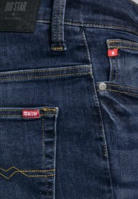 Zbliżenie na ciemnoniebieskie dżinsy z denimu, pokazujące tylne kieszenie, żółte przeszycia, metalowy nit oraz czerwone metki z białą gwiazdą i napisem "BIG STAR".