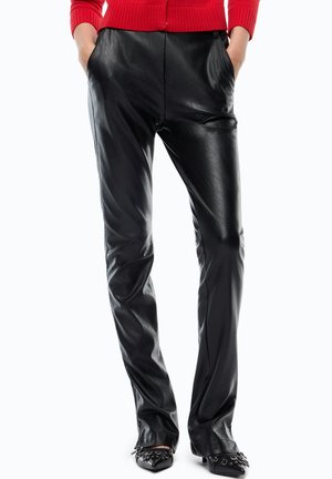 Persona che indossa pantaloni in pelle nera, maglione rosso e scarpe nere con punta appuntita e borchie metalliche, mani in tasca, in piedi su sfondo bianco.