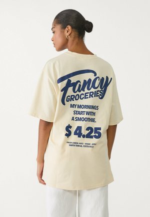 Cremefarbenes, übergroßes T-Shirt mit einem dunkelblauen Grafikdruck auf der Rückseite, der Informationen über Smoothies und Preise enthält. Kurzärmlig, lockere Passform.