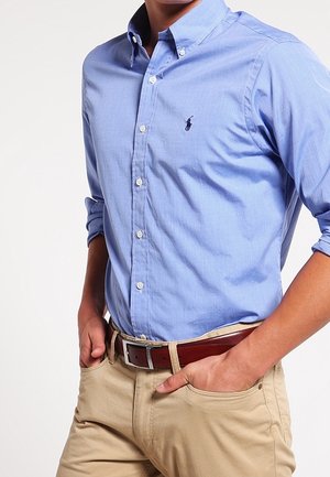 Mężczyzna w jasnoniebieskiej koszuli na guziki z podwiniętymi rękawami, beżowych spodniach i brązowym skórzanym pasku, z rękami w kieszeniach.