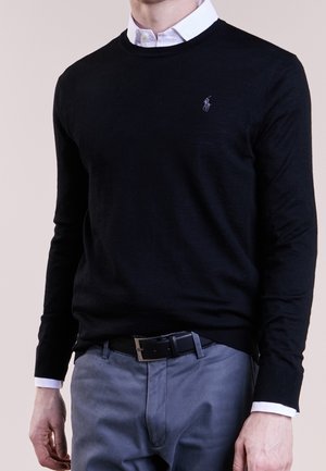 Mand iført sort langærmet sweater over hvid skjorte med krave, lysegrå bukser og sort bælte, stående foran en neutral baggrund.