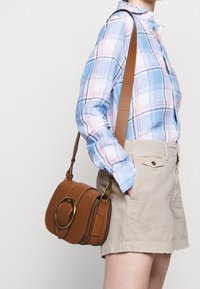 Žena v béžových šortkách a modré kostkované košili nesoucí hnědou koženou tašku přes rameno se zlatou sponou.