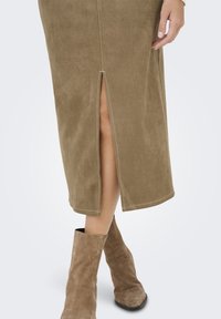Falda midi de ante marrón con una abertura frontal. Combinada con botas de ante marrón claro hasta el tobillo. Costuras visibles y textura suave.