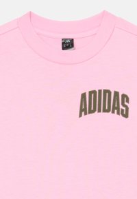 Camiseta de algodón rosa con escote redondo y mangas cortas, que presenta un logo verde en arco de "ADIDAS" en el lado izquierdo.
