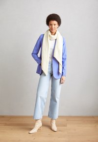 Femme debout sur un sol en bois portant un blazer bleu clair, une écharpe blanche, un haut blanc, un jean bleu clair et des bottines beiges devant un mur gris uni.