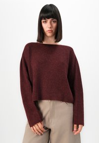 Jeune femme aux cheveux noirs raides, portant un pull ample bordeaux et un pantalon beige, se tenant devant un fond blanc uni.