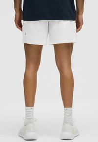 Shorts atléticos blancos con una textura suave, combinados con zapatillas blancas y calcetines de color claro. Vista desde atrás, mostrando las piernas y el calzado.