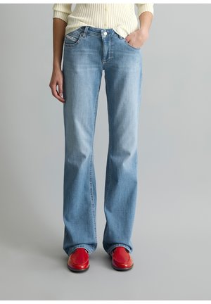 Person trägt hellblaue Jeans mit geradem Bein, einen cremefarbenen Rippstrickpullover, die Hände in den Taschen und sieht rote Schuhe.