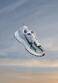 Die Nike-Sneaker verfügen über ein graues Mesh-Obermaterial, dunkle blaue Akzente, eine Schnürung, eine dicke weiße Sohle und ein markantes strukturiertes Design.