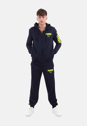 Giovane uomo in piedi con le mani nelle tasche che indossa una tuta Puma blu navy con loghi giallo neon e sneakers bianche su sfondo uniforme.